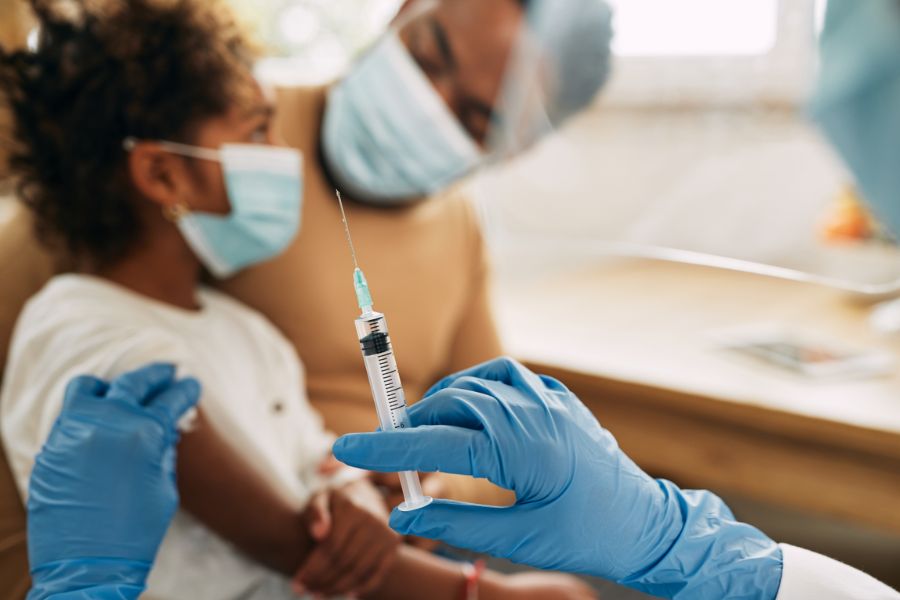 Criança usando máscara sendo preparada para vacinação por profissional de saúde